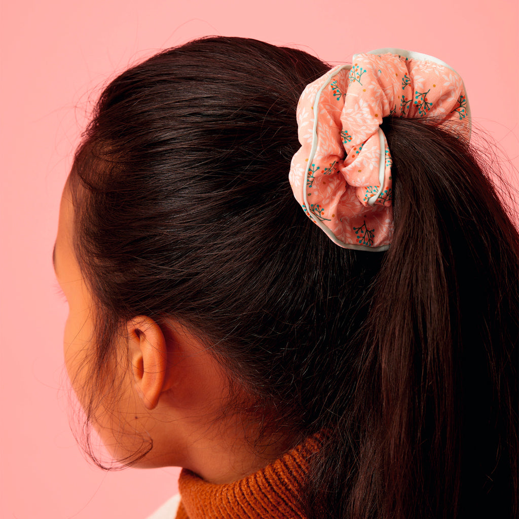 A person with dark hair is turned away, showcasing a pink floral scrunchie. The background is a pink hue, complementing the cozy autumn vibe.