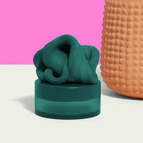 Dark-green focus putty on its container, set against a pink background next to a textured terracotta vase.