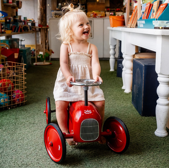 Child laughing while riding a red Vilac ride-on toy car in a toy shop, surrounded by shelves and colourful baskets of toys