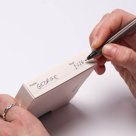 A person writing "GEORGE" and "2026" on the side of a small white box with a pen.