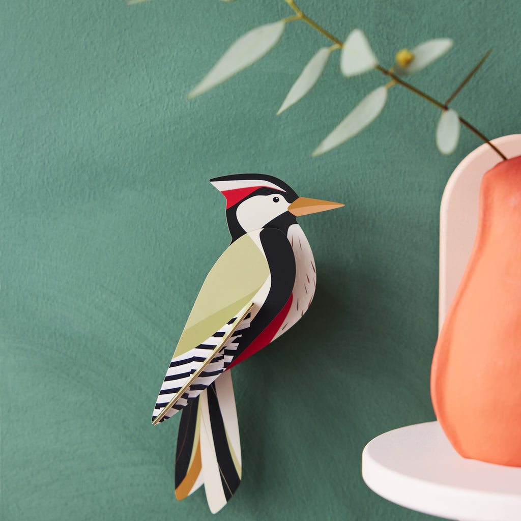 A colorful paper bird with a red, black, and white pattern is mounted on a green wall. Nearby is a branch and the edge of an orange vase, creating a whimsical scene.