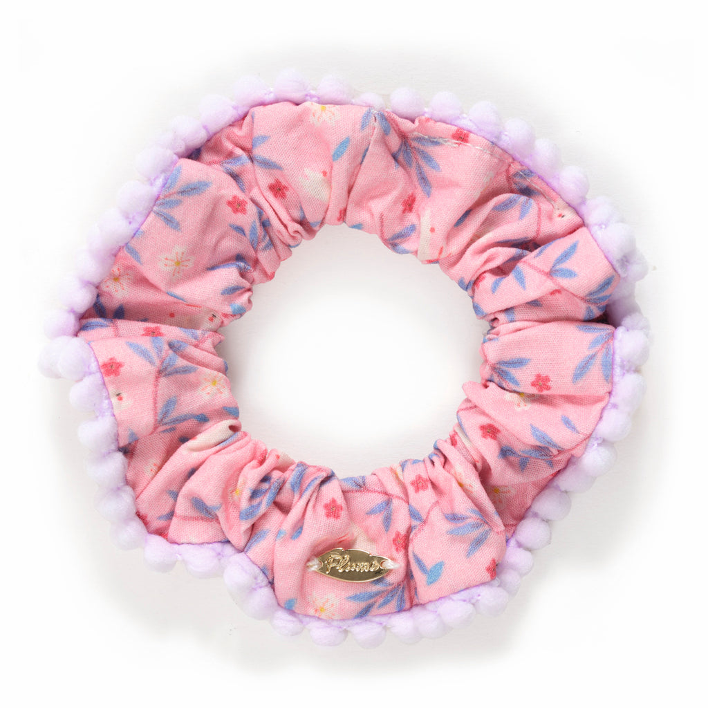 Pink floral scrunchie with pom-pom edge and a small, gold metallic tag. Light and playful vibe with a soft, textured design.