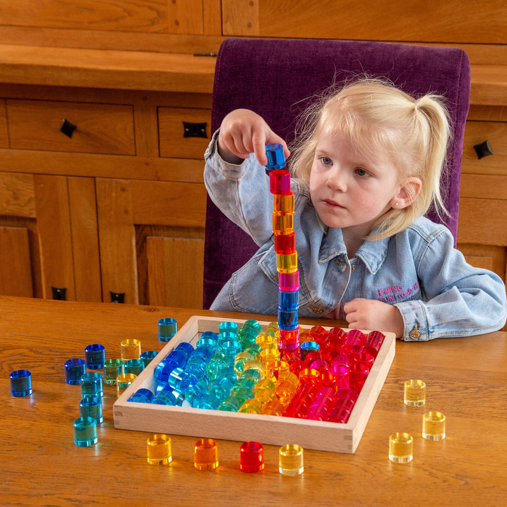 Young girl stacking colorful translucent discs in a wooden tray, sitting at a wooden table. She is focused and wears a light denim jacket.