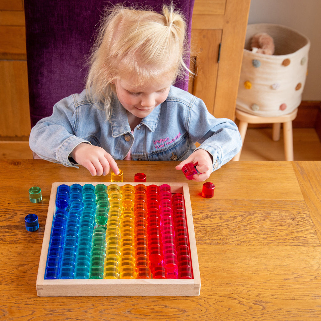 Young girl stacking colorful translucent discs in a wooden tray, sitting at a wooden table. She is focused and wears a light denim jacket.