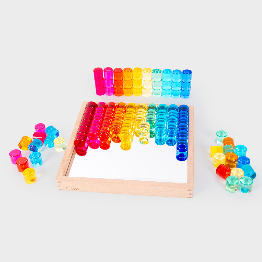 Colorful transparent counters in red, orange, yellow, green, and blue, arranged neatly on a mirrored wooden tray and scattered around it, creating a playful and vibrant tone.