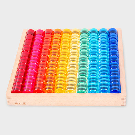 A wooden tray holds colorful transparent counters arranged in a vibrant rainbow pattern from red to blue, creating a bright and cheerful display.