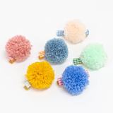 Six colorful pom-pom hair clips arranged on a white background with gingham ribbon bases.