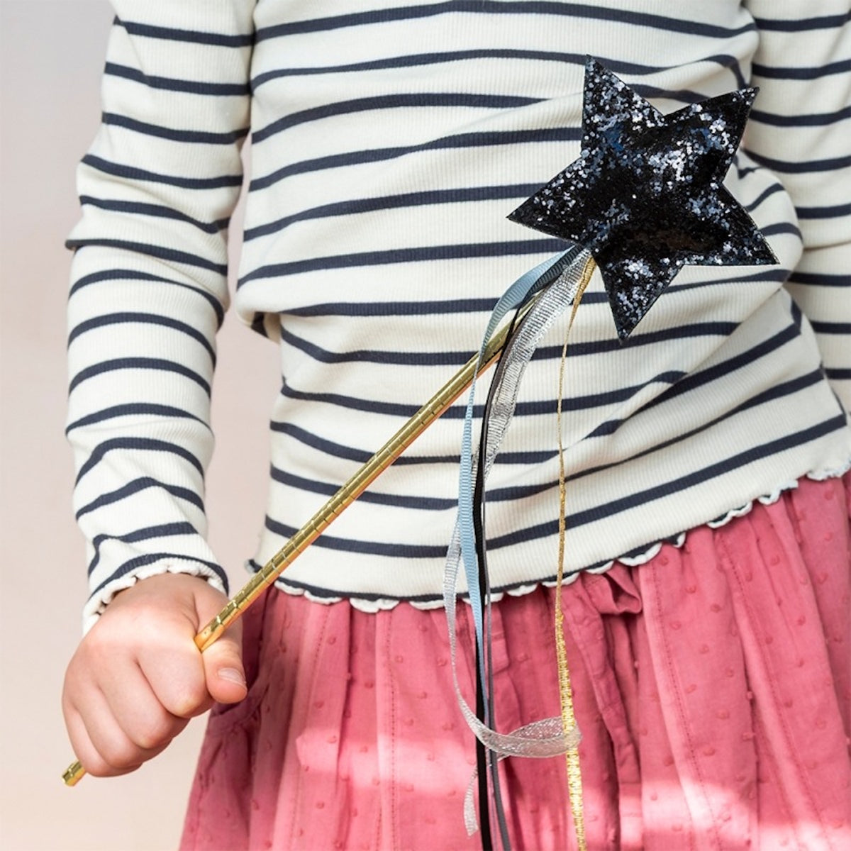 Child in striped shirt and pink skirt holding a black glittery star magic wand with ribbons.