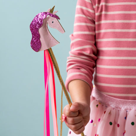 A child holding a pink unicorn wand with a glittery purple mane, gold stick, and hanging ribbons.