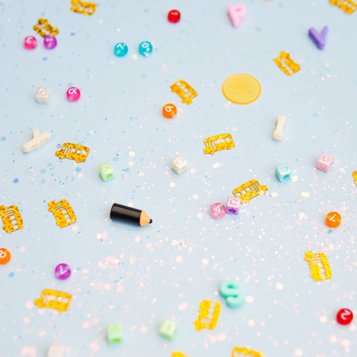 Colorful letter beads, number beads, pencil charm, and yellow school bus confetti scattered on light blue glitter.