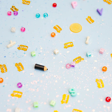 Colorful letter beads, number beads, pencil charm, and yellow school bus confetti scattered on light blue glitter.