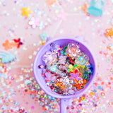 A purple cup filled with fairy-themed trinkets, flowers, and glitter against a sparkly pink background.