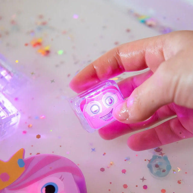 Hand holding a glowing pink light-up sensory cube with a cute face above glittery water.