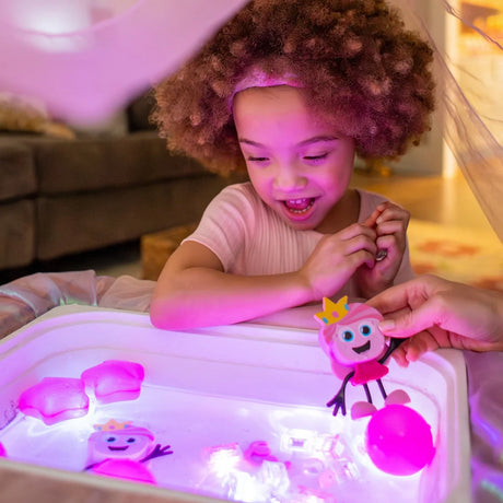 Young girl smiling while playing with glowing pink sensory cubes and character figures in a water tray.