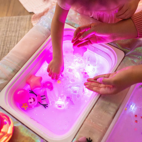 Child and adult hands playing with glowing pink sensory light cubes and toys in a water tray.