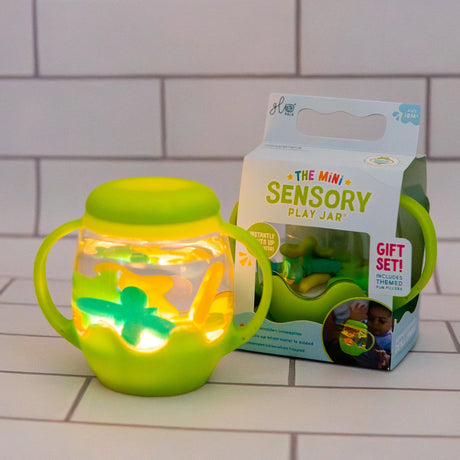 Glowing green Glo Pals Mini Sensory Play Jar with floating shapes, set next to its retail box.
