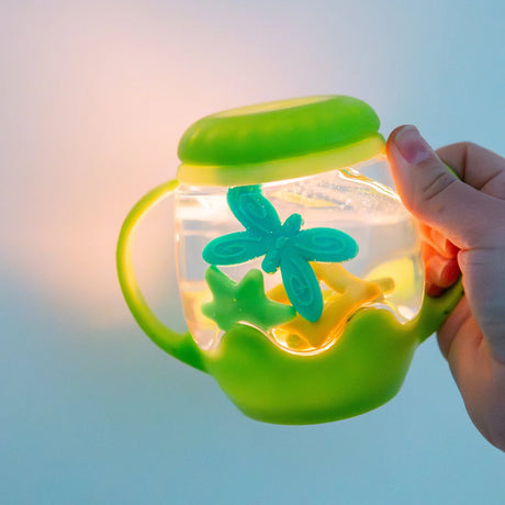 A hand holds a glowing green sensory jar with handles, filled with floating star and dragonfly shapes.