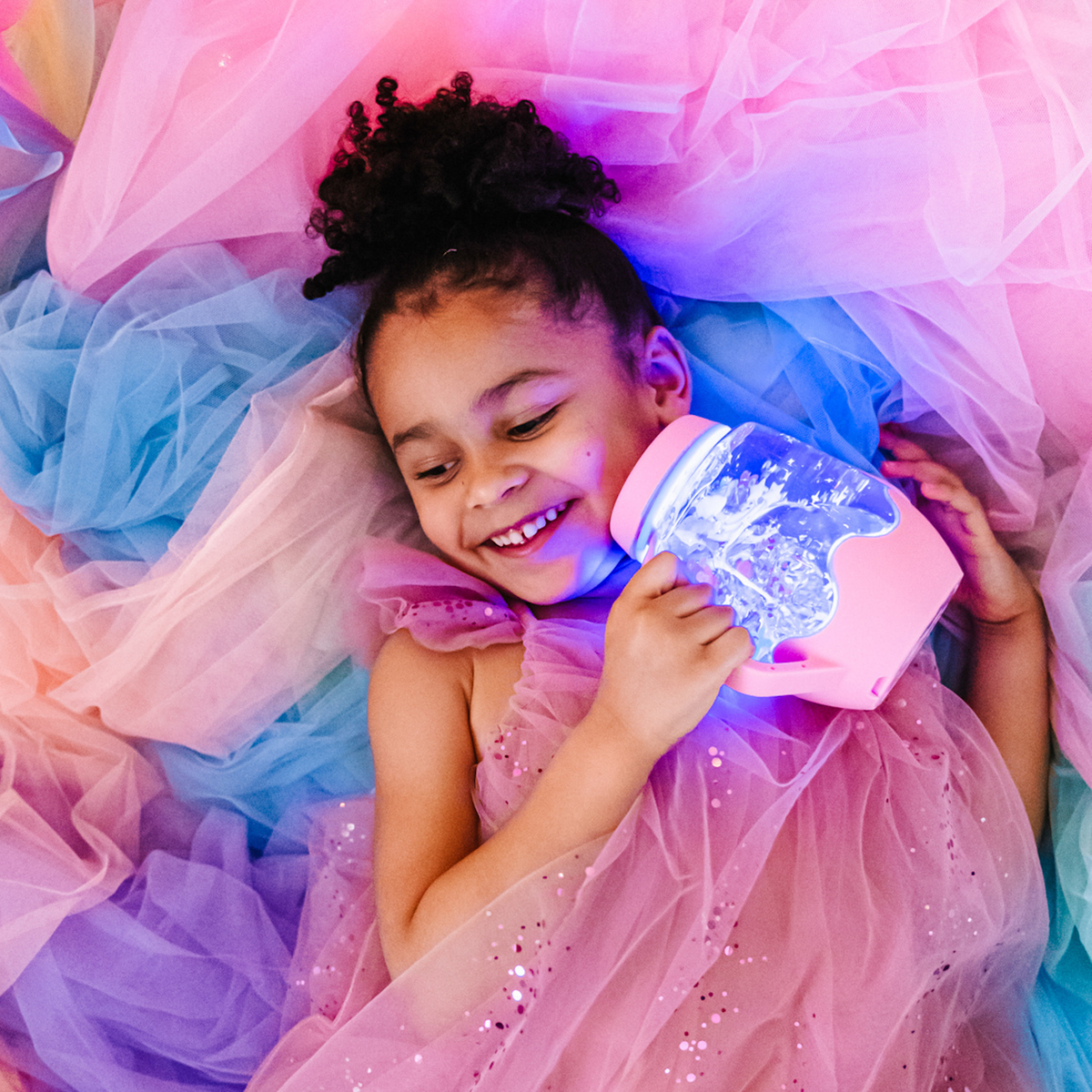 Girl in a pink dress hugging a glowing light-up sensory jar against colorful tulle fabric.