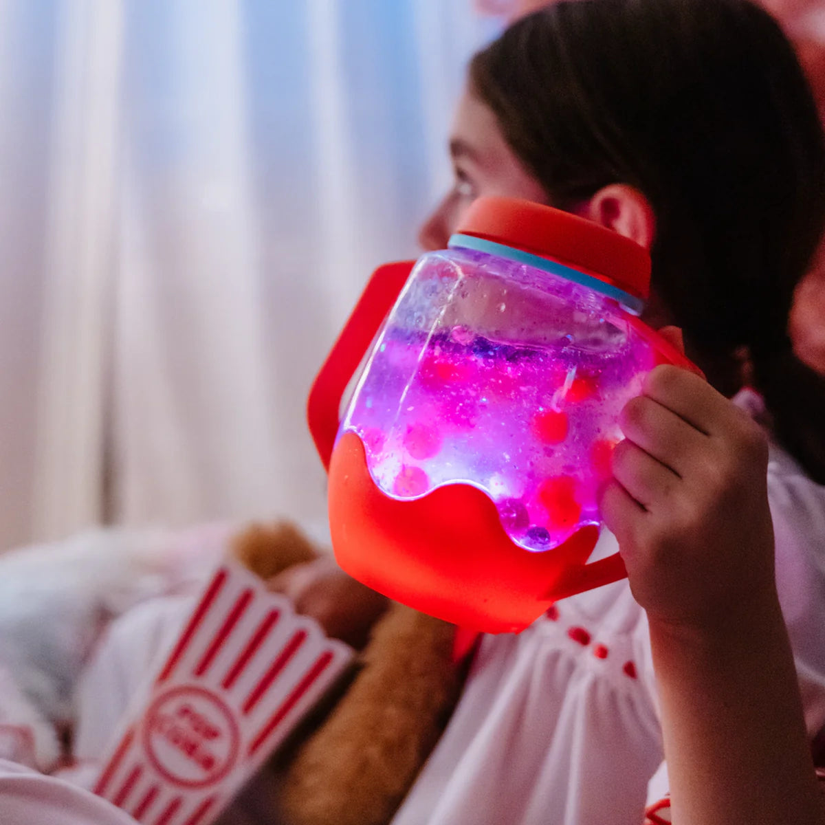 Child holding a glowing pink and purple light-up sensory jar with red handles during a cozy movie night.