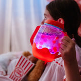 Child holding a glowing pink and purple light-up sensory jar with red handles during a cozy movie night.