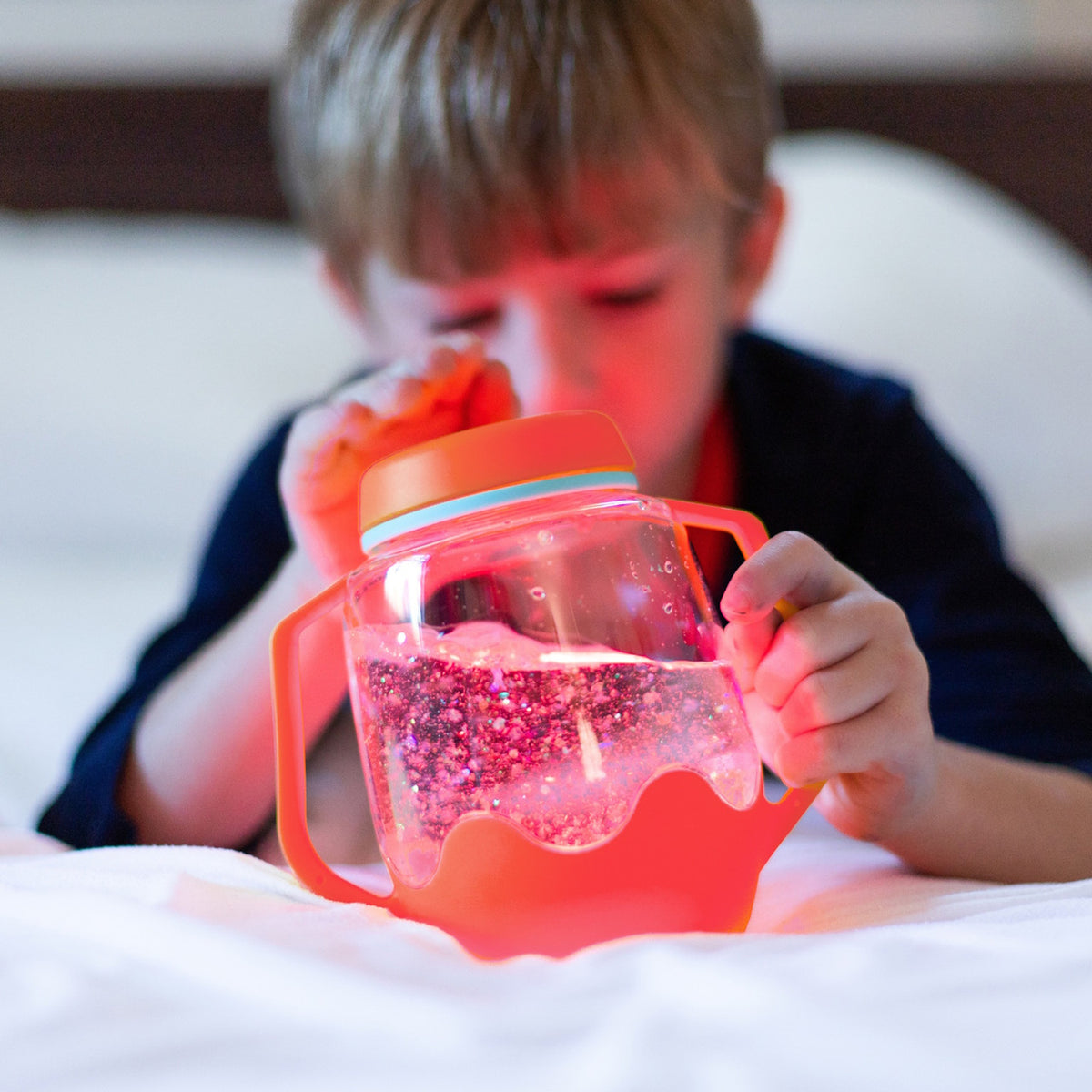 Young boy playing with a glowing orange sensory jar filled with swirling glitter and liquid.