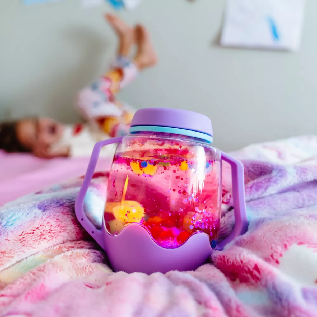 Glowing pink Glo Pals sensory play jar with a purple base resting on a blanket, with a child blurred in the background.