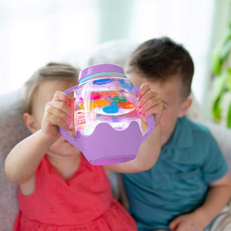 Two children holding up a glowing purple Glo Pals sensory play jar filled with water and colorful floating pieces.
