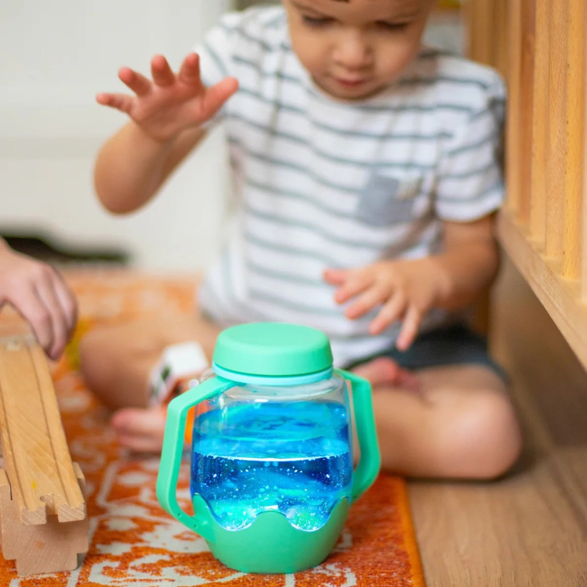 Toddler sitting on the floor playing near a light-up teal sensory jar filled with glowing blue liquid.