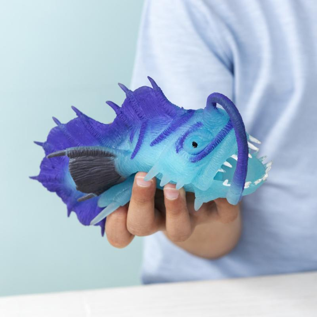 A child holds a vibrant blue and purple toy anglerfish with pronounced teeth and fins, creating a playful and curious atmosphere against a light background.