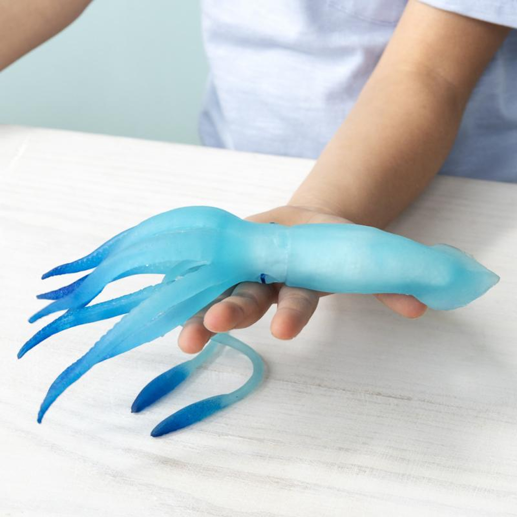 Child's hand holding a blue rubber squid toy with gradient color from light blue to dark blue on tentacles, placed on a light wooden surface.