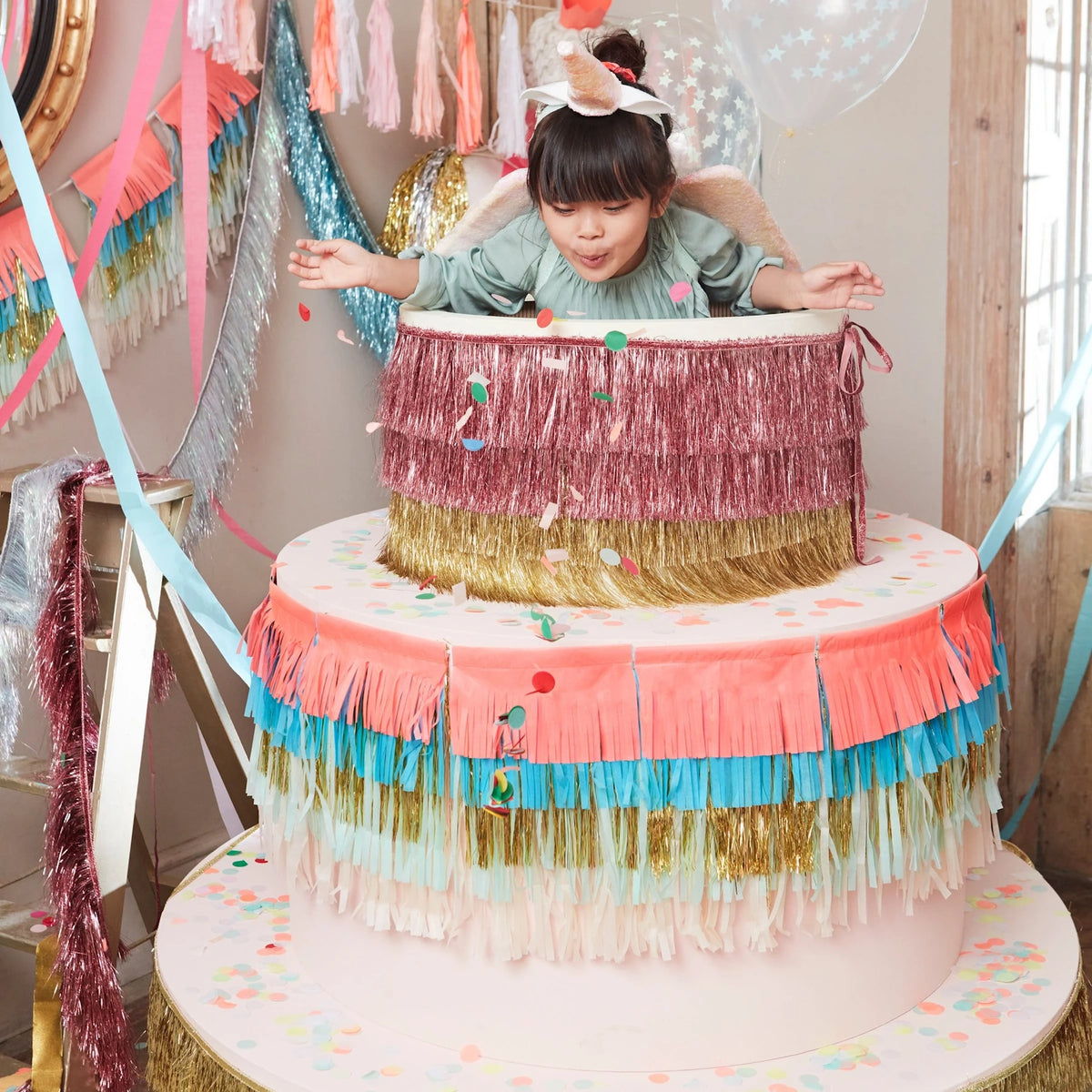 Young girl popping out of a oversized fringe-decorated cake prop surrounded by party decorations.