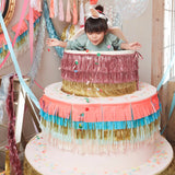 Young girl popping out of a oversized fringe-decorated cake prop surrounded by party decorations.