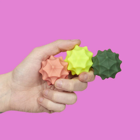 A hand holding three connected spiky silicone magnetic fidget spheres on a pink background.