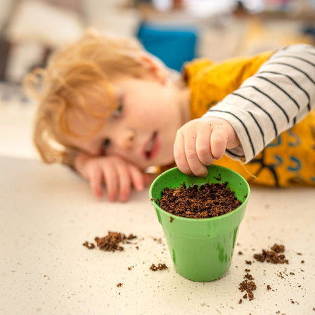 A blond child touches the soil in a small green pot, with dirt scattered on the surface.