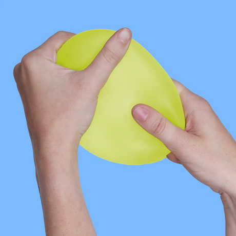 Two hands squeezing a yellow stress ball against a solid blue background. The hands are positioned gently, conveying a sense of relaxation.