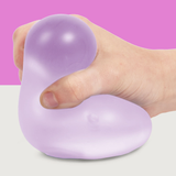 A hand squeezes a soft, purple stress ball against a two-tone background, with pink on top and white below. The scene conveys relaxation and calm.