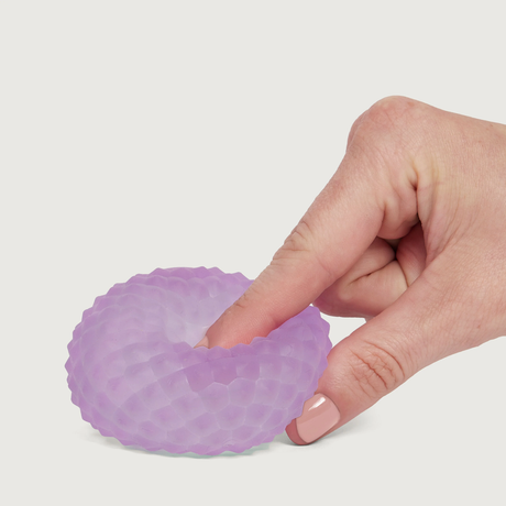 A hand with light skin gently presses a textured, purple, donut-shaped stress ball on a white background, conveying a sense of calm and relaxation.