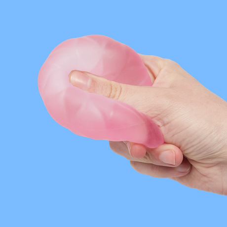 A hand squeezing a pink, textured stress ball against a blue background. The ball is being compressed, illustrating flexibility and stress relief.