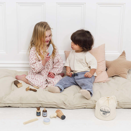 Two young children sitting on a cushion, playing with a wooden hairdresser and barber toy set.