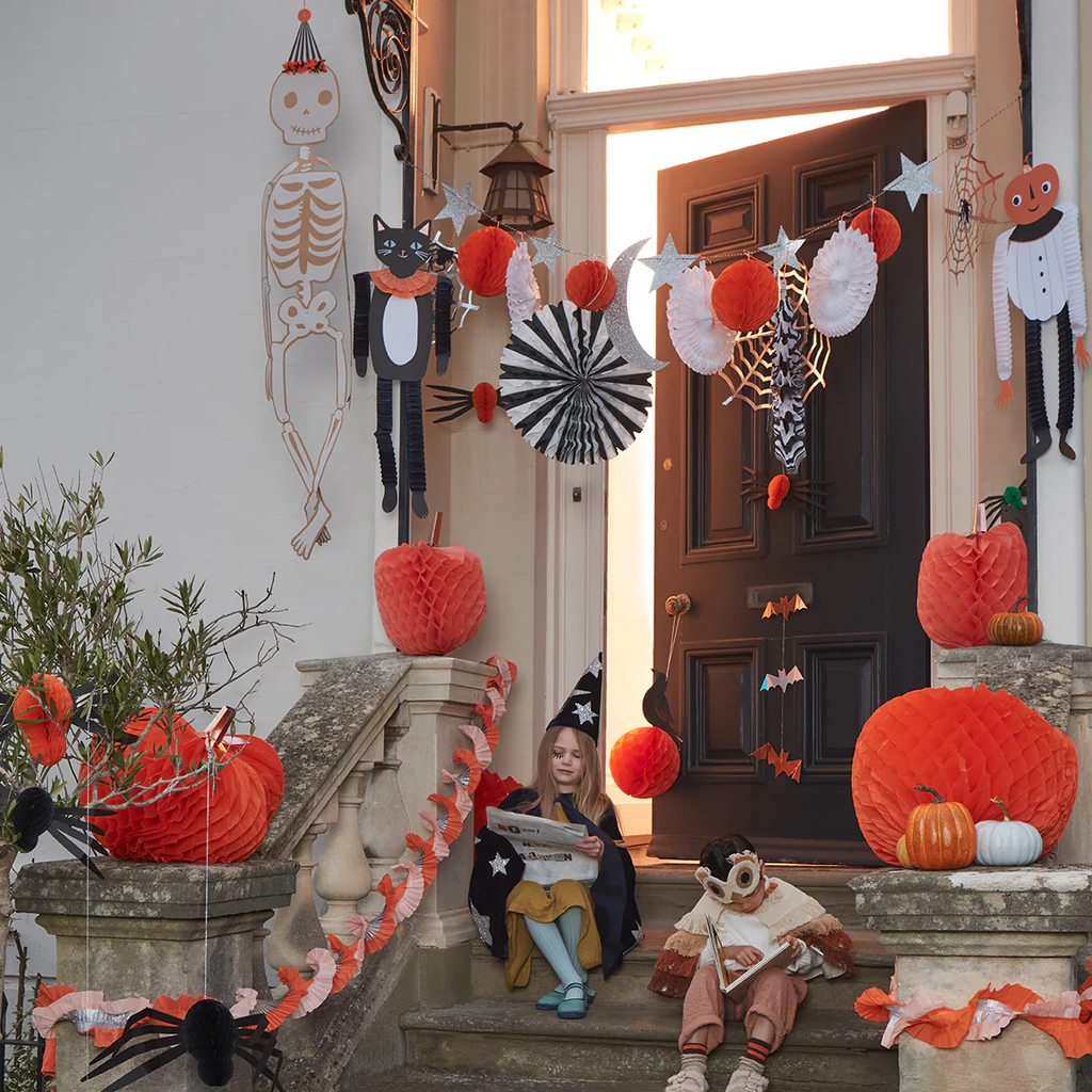 Festive Halloween porch with a girl in a witch hat reading on the steps. Decor includes pumpkins, skeletons, black cats, and garlands in orange, black, and white.