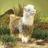 Alpaca hand puppet standing outdoors among rocks and grassy foliage.