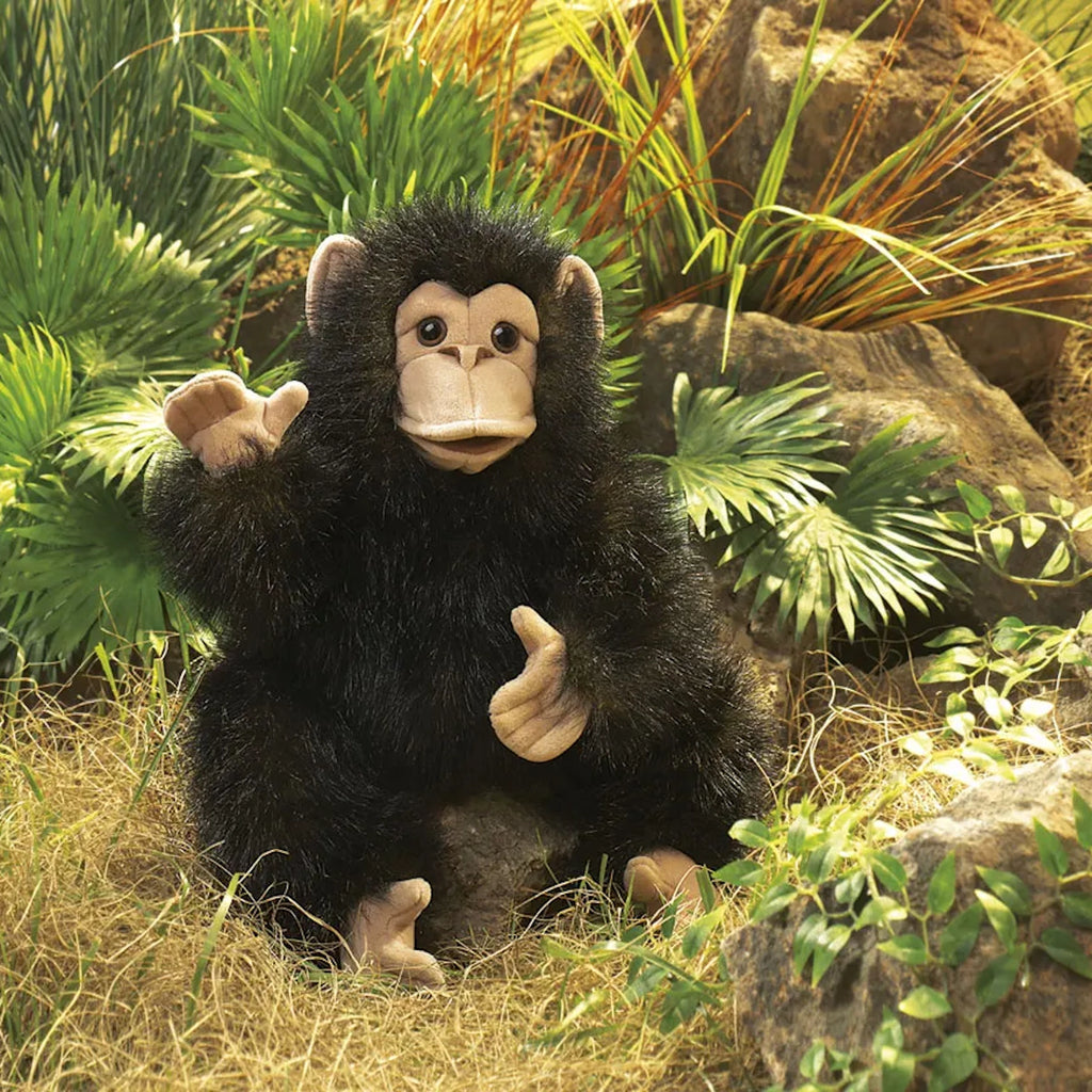 A stuffed monkey puppet with black fur and beige face sits in lush greenery, surrounded by rocks and plants, giving a playful and lively tone.