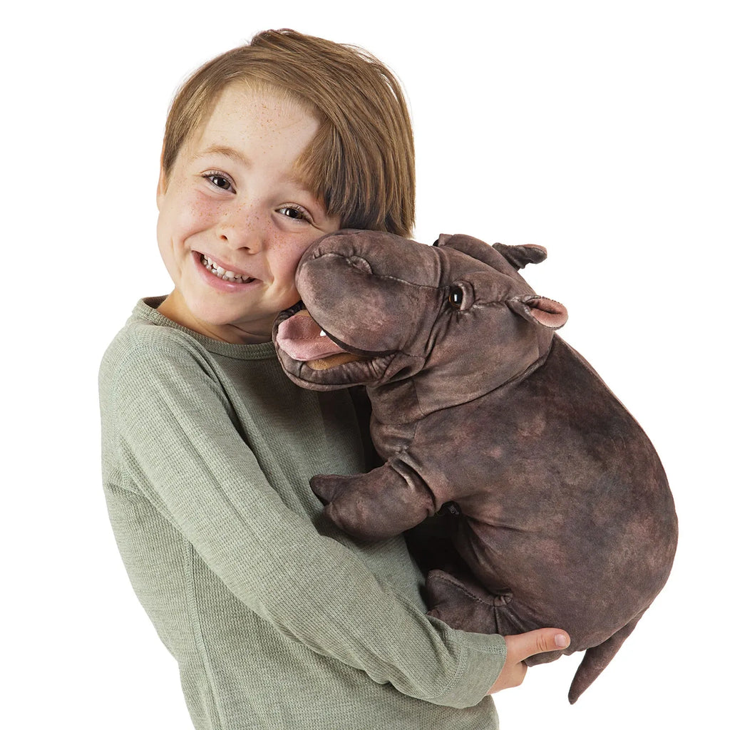 A smiling child with light brown hair embraces a large, plush hippopotamus toy. The child wears a light green sweater and looks joyful.