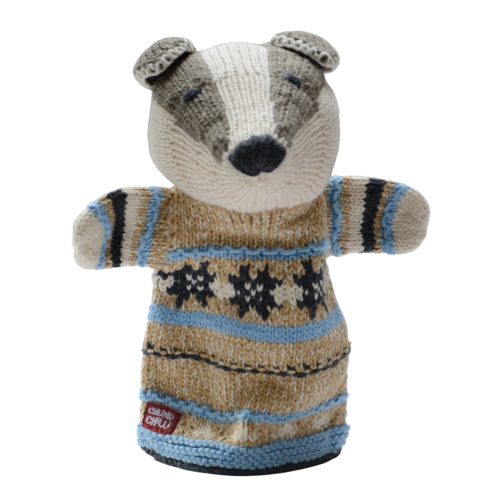 A knitted badger puppet wearing a cozy, patterned sweater in beige, blue, and black. It has a friendly expression and is set against a white background.