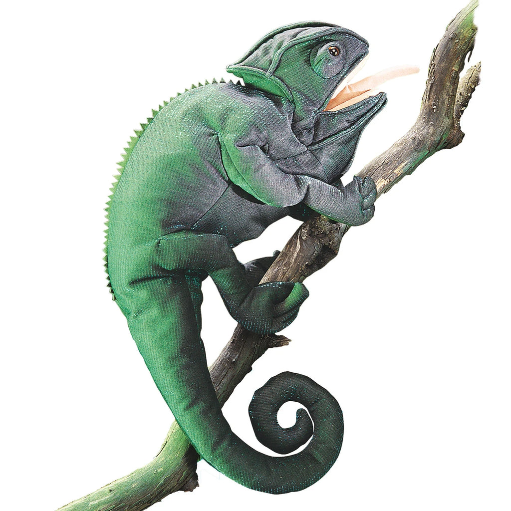 A green chameleon puppet, with textured skin and a curled tail, clings to a wooden branch. Its mouth is open, creating a playful and lively tone.