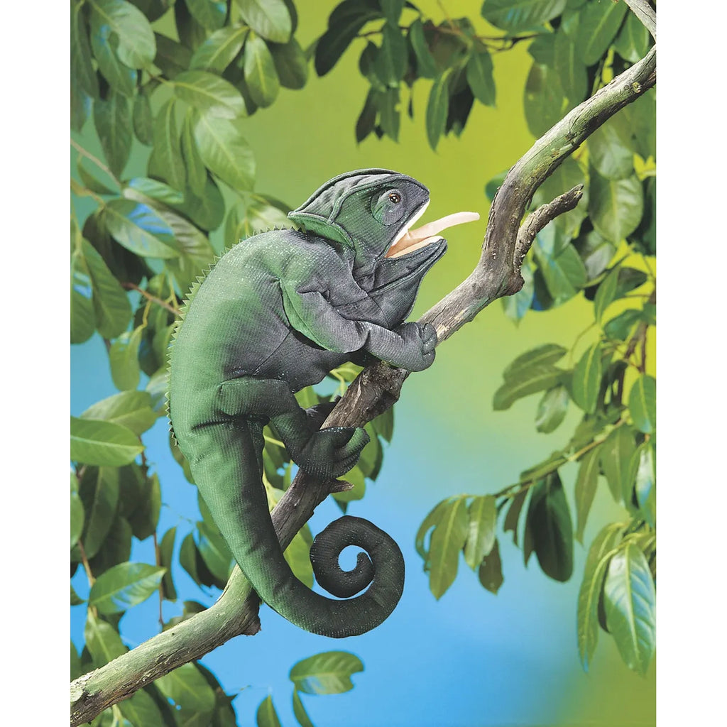 A green chameleon sits on a tree branch with its mouth open and tongue extended. Surrounding leaves create a vibrant backdrop of greens and blues.
