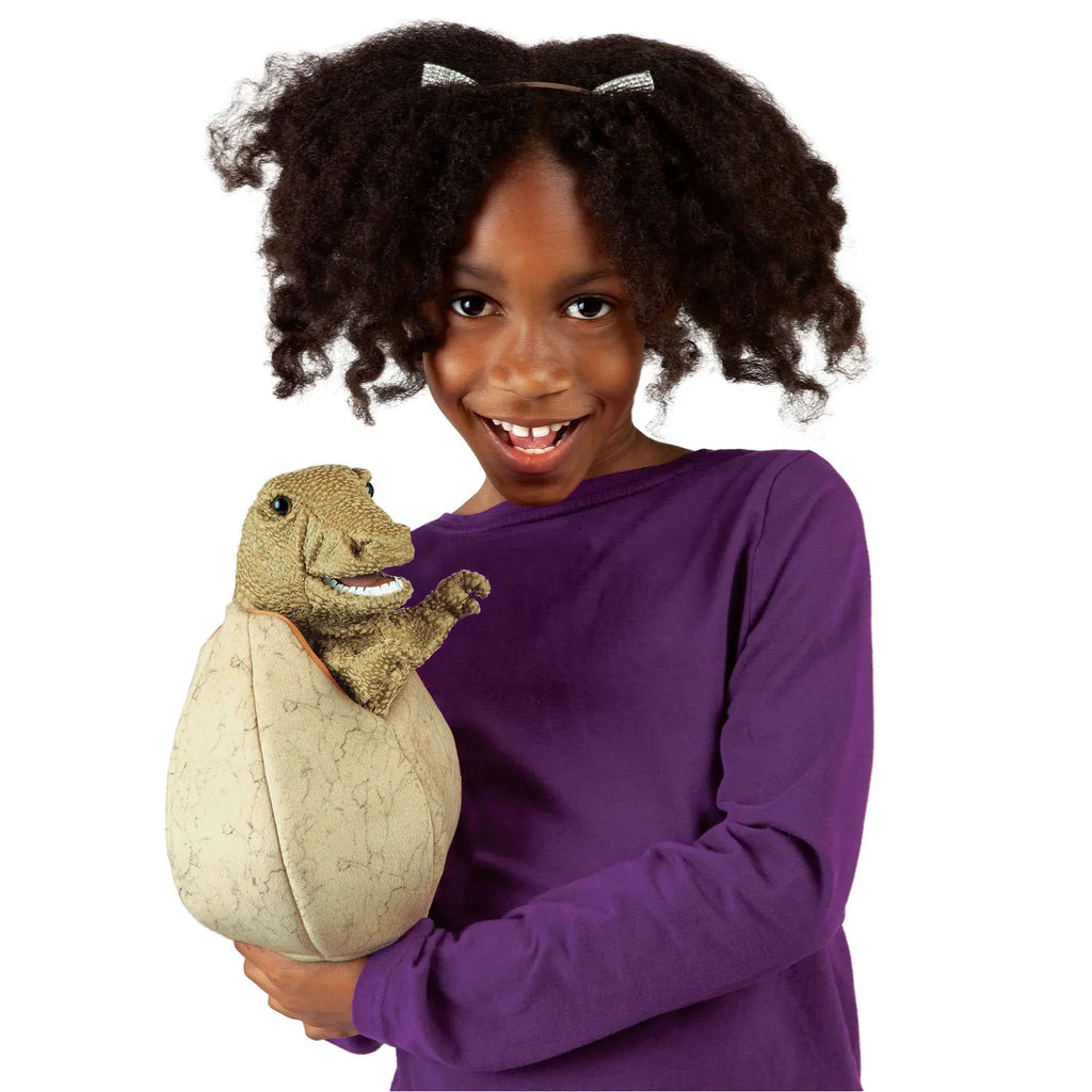 A joyful child with curly hair and a purple top smiles widely, holding a dinosaur puppet emerging from an egg. The scene conveys excitement and playfulness.