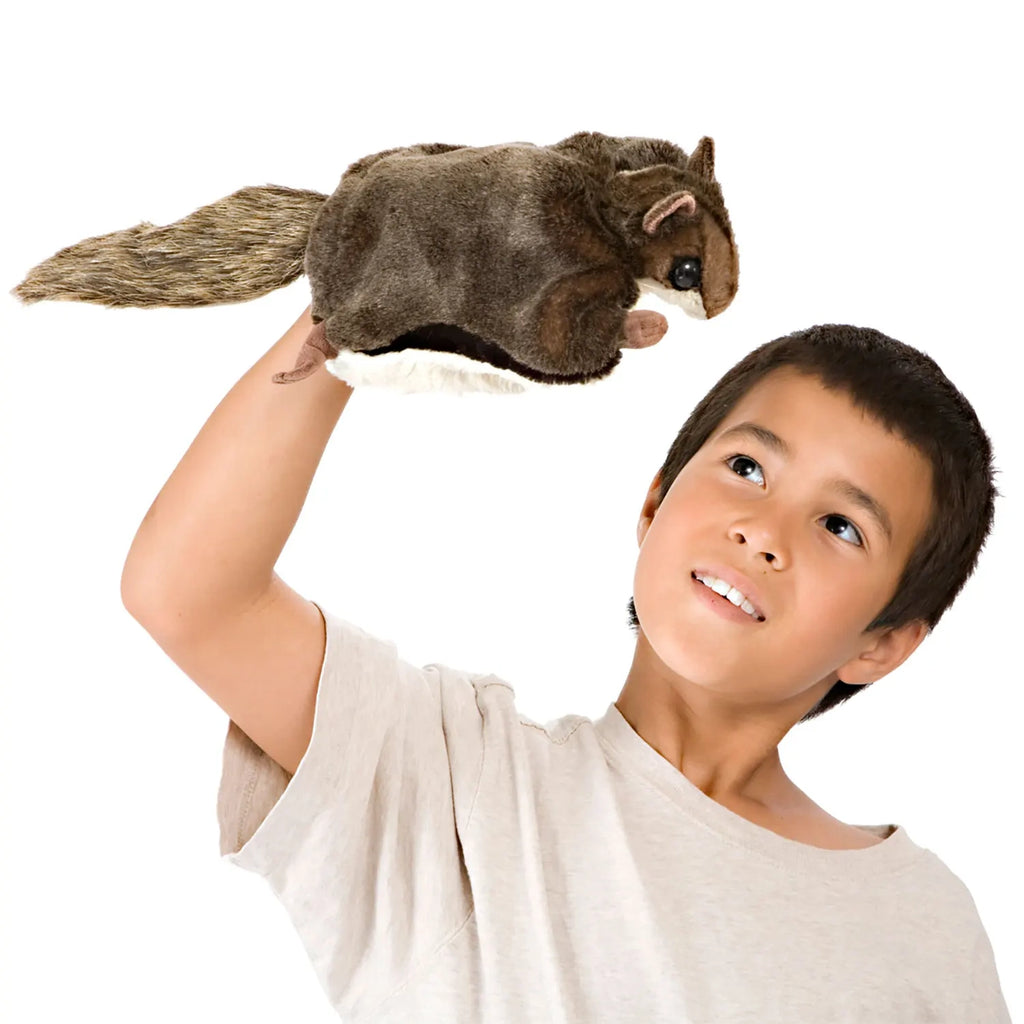 A child wearing a light beige shirt holds a plush squirrel puppet on their hand, looking up at it with a joyful expression. The background is white.