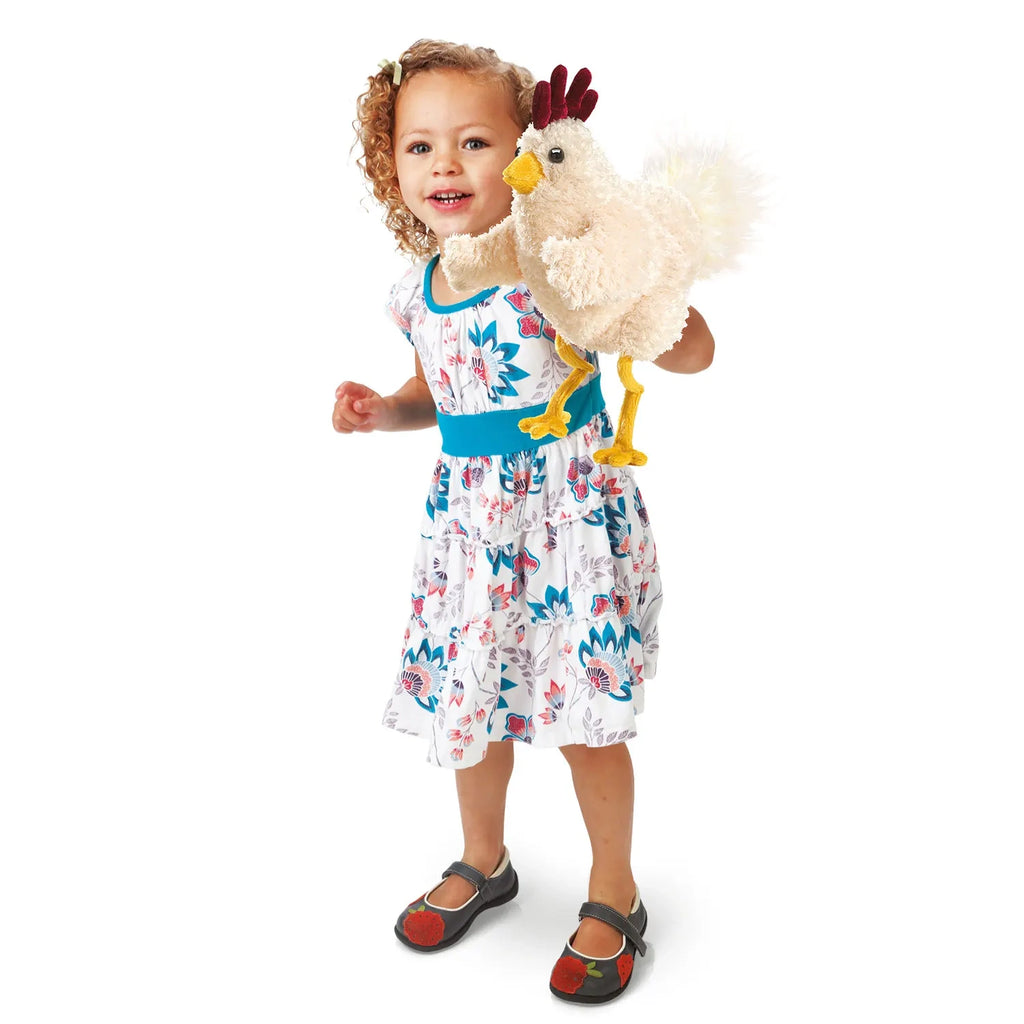 A smiling child with curly hair holds a plush chicken toy. They're wearing a floral dress with a blue waistband and black shoes on a white background.