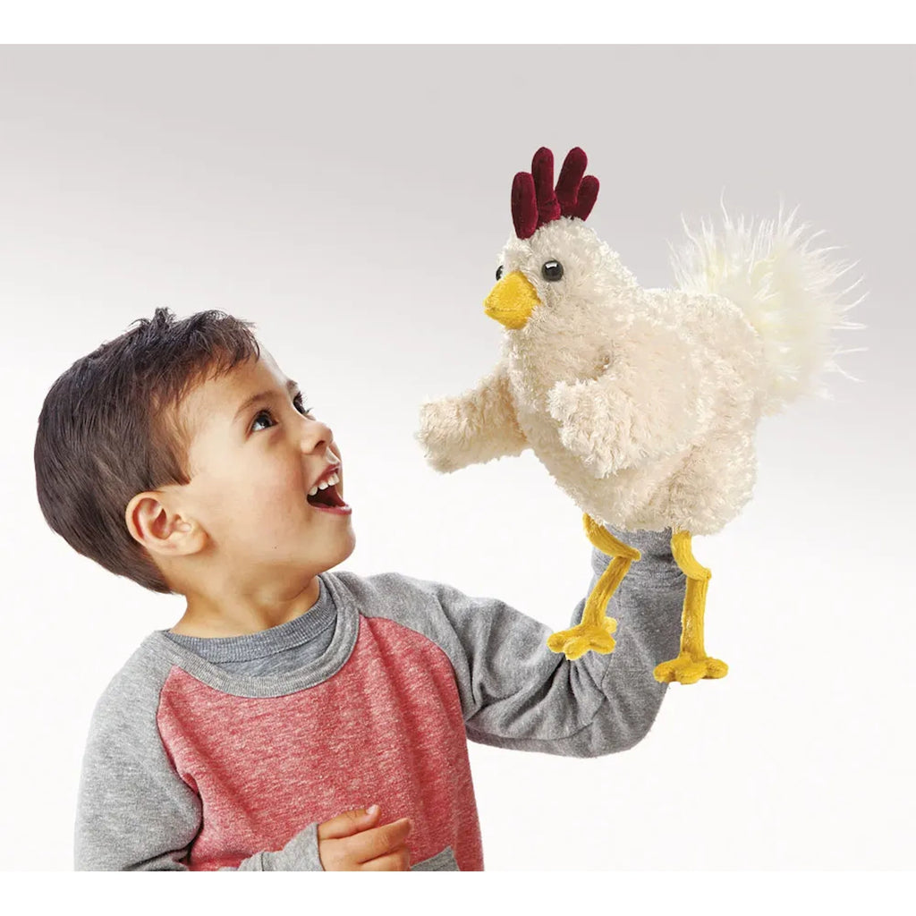A smiling child in a red and gray sweater holds up a fluffy white chicken puppet with yellow beak and feet. The child looks excited and engaged.